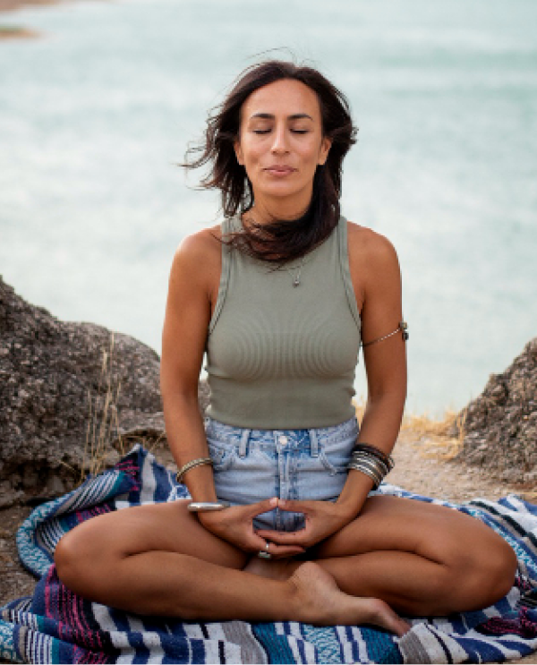 Woman meditating cross-legged on a woven blanket by the water, eyes closed, hands resting in her lap