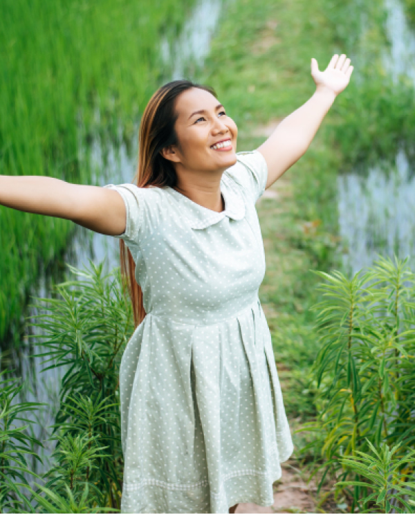 Smiling woman in a polka-dot dress with arms spread wide on a path between green rice fields