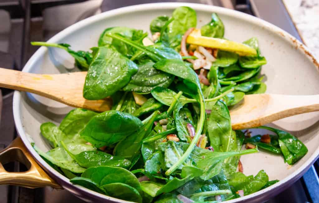 Fresh spinach tossed with red onion and lemon in a skillet with two wooden spoons