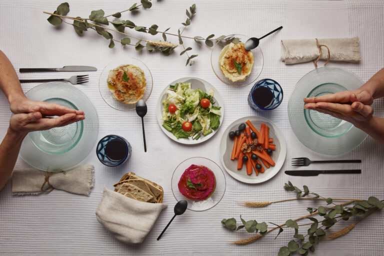 Overhead table of salad, hummus, beet dip and carrots, with two people's hands held palms together