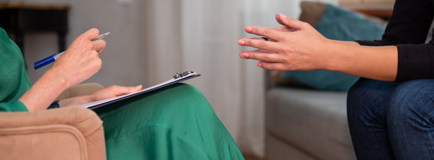 Therapist with a clipboard and pen listening to a client gesturing with clasped hands in a counseling session