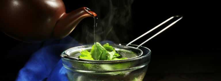Hot water poured from a clay teapot over fresh green leaves in a strainer set in a glass bowl