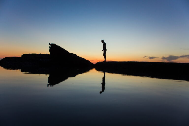 Silhouette of a person standing on rocks at sunset, reflected in still water