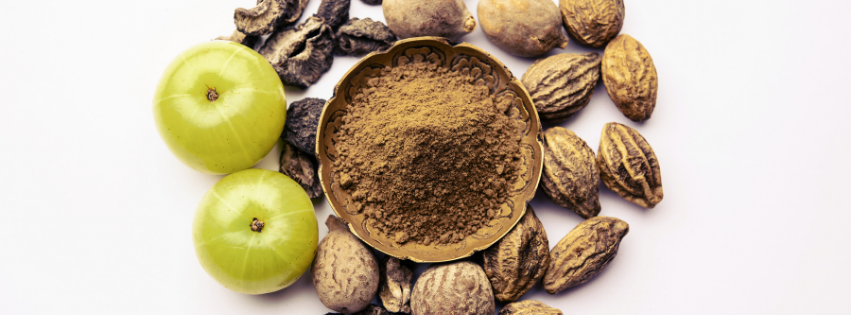 Bowl of brown Triphala powder surrounded by fresh amla, dried haritaki and bibhitaki fruits